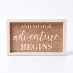 A rectangular wooden sign with a light painted frame and a medium brown wood-grain center panel. The text reads 'AND SO OUR ADVENTURE BEGINS' in cream-colored lettering, with 'adventure' in a cursive script font.