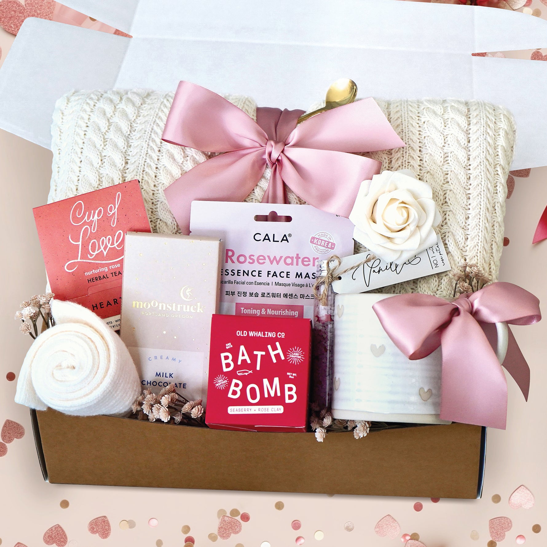 Be My Valentine gift box comes with cream cable knit blanket tied with a blush ribbon, rose face mask, Seaberry and Rose Bath Bomb, milk chocolate moonstruck bar, cream knit socks, cup of love herbal tea, vanilla rose bath salt tube, ceramic hearts mug, gold demitasse spoon, and faux cream rose head
