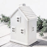 A white ceramic house with window cutouts, photographed on a light wood surface with soft greenery in the background.