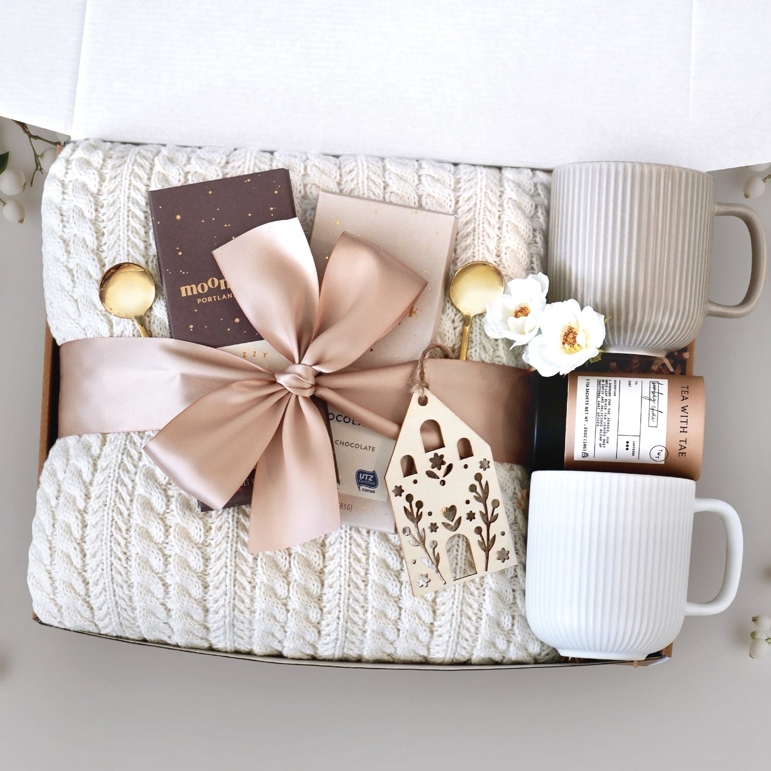 Cozy Home Blanket Box comes with a cream cable knit blanket tied with a tan ribbon, one Moonstruck Milk Chocolate Bar, one Moonstruck Dark Chocolate Espresso Bar, one Nordic wooden house ornament, one Tea with Tae Tea Tube, one cream ribbed mug, one grey ribbed mug, and two gold demitasse spoons