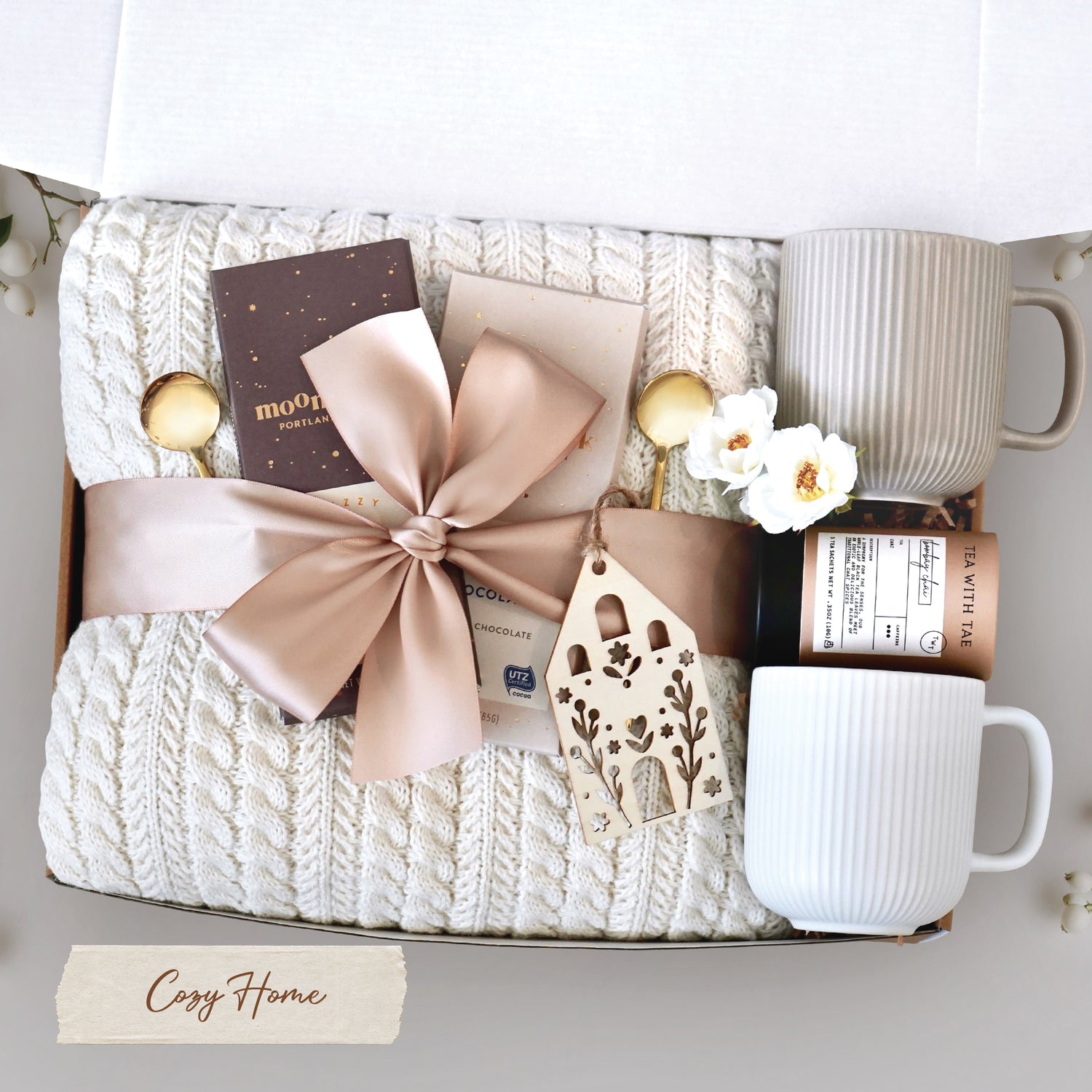 Cozy Home Blanket Box comes with a cream cable knit blanket tied with a tan ribbon, one Moonstruck Milk Chocolate Bar, one Moonstruck Dark Chocolate Espresso Bar, one Nordic wooden house ornament, one Tea with Tae Tea Tube, one cream ribbed mug, one grey ribbed mug, and two gold demitasse spoons