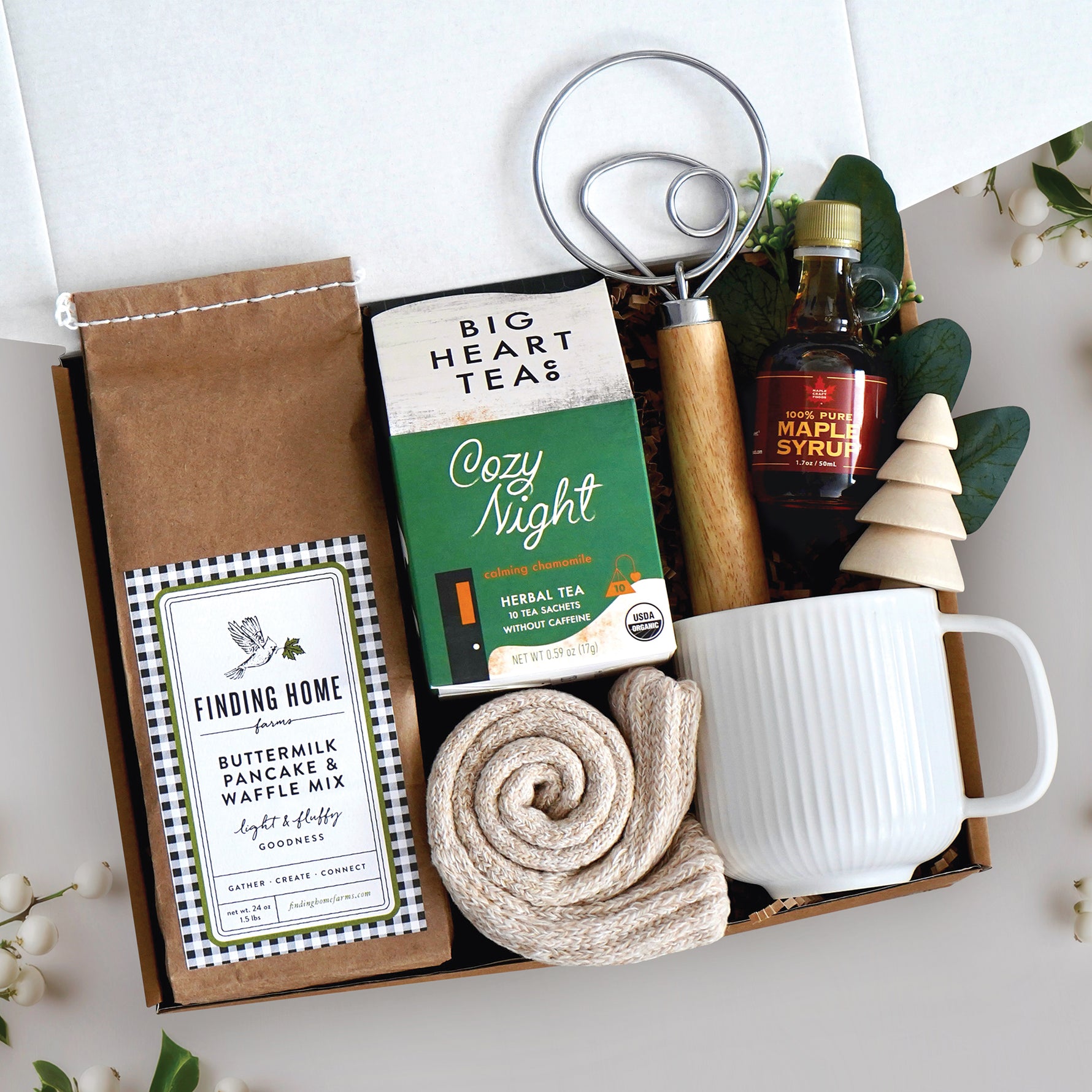 Cozy Kitchen Gift Box come with a ribbed white ceramic mug, dough whisk, 24oz pancake mix, cozy night tea bags 10 count, cozy boot socks, 1.7ox maple syrup, and faux eucalyptus sprig