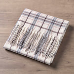 A folded cream plaid scarf with fringe, displayed on a light brown wooden surface. The scarf features a cream base with a classic plaid pattern in coordinating shades of dark brown, dusty blue, and rust.