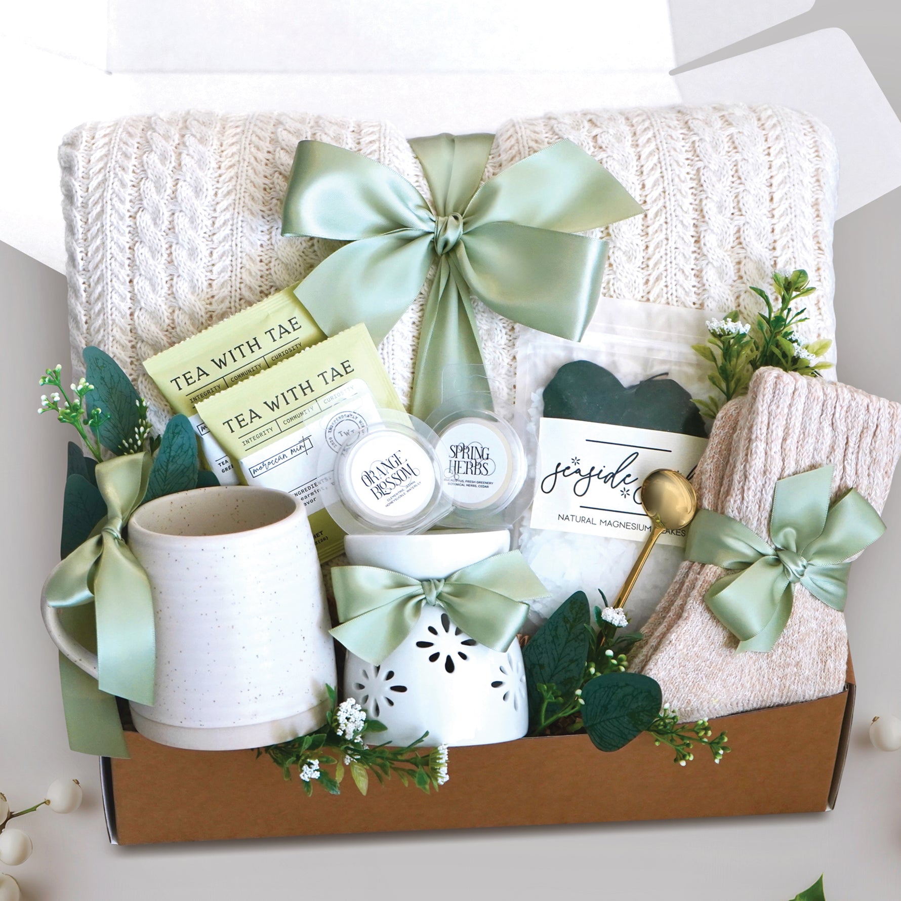 The Keep on Growing Gift Box xome with a cream cable knit blanket tied with a sage ribbon, Wildflower Pop Beehive Mug, Starburst wax warmer and 2 wax melts, 2 tea bags, Seaside Soak bath salts, cozy boot socks, and gold demitasse spoon