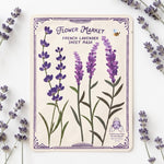 A Flower Market French Lavender Sheet Mask with illustrated purple lavender flowers, green stems, and a small bee on an off-white background, surrounded by real lavender sprigs on a white surface.