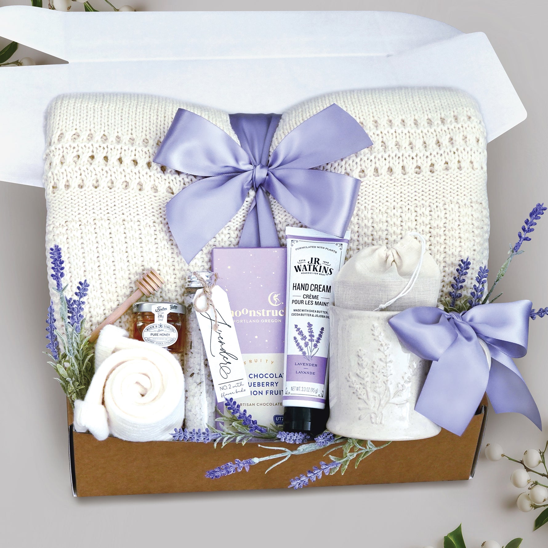 Lovely Lavender comes with a cream lace knit blanket tied with a lavender ribbon, cream knit socks, lavender ceramic mug, 2 tea bags, moonstruck dark chocolate blueberry and passionfruit bar, JR Watkins Lavender hand cream, tube of floral bath salts, mini honey and dipper, and faux lavender sprig