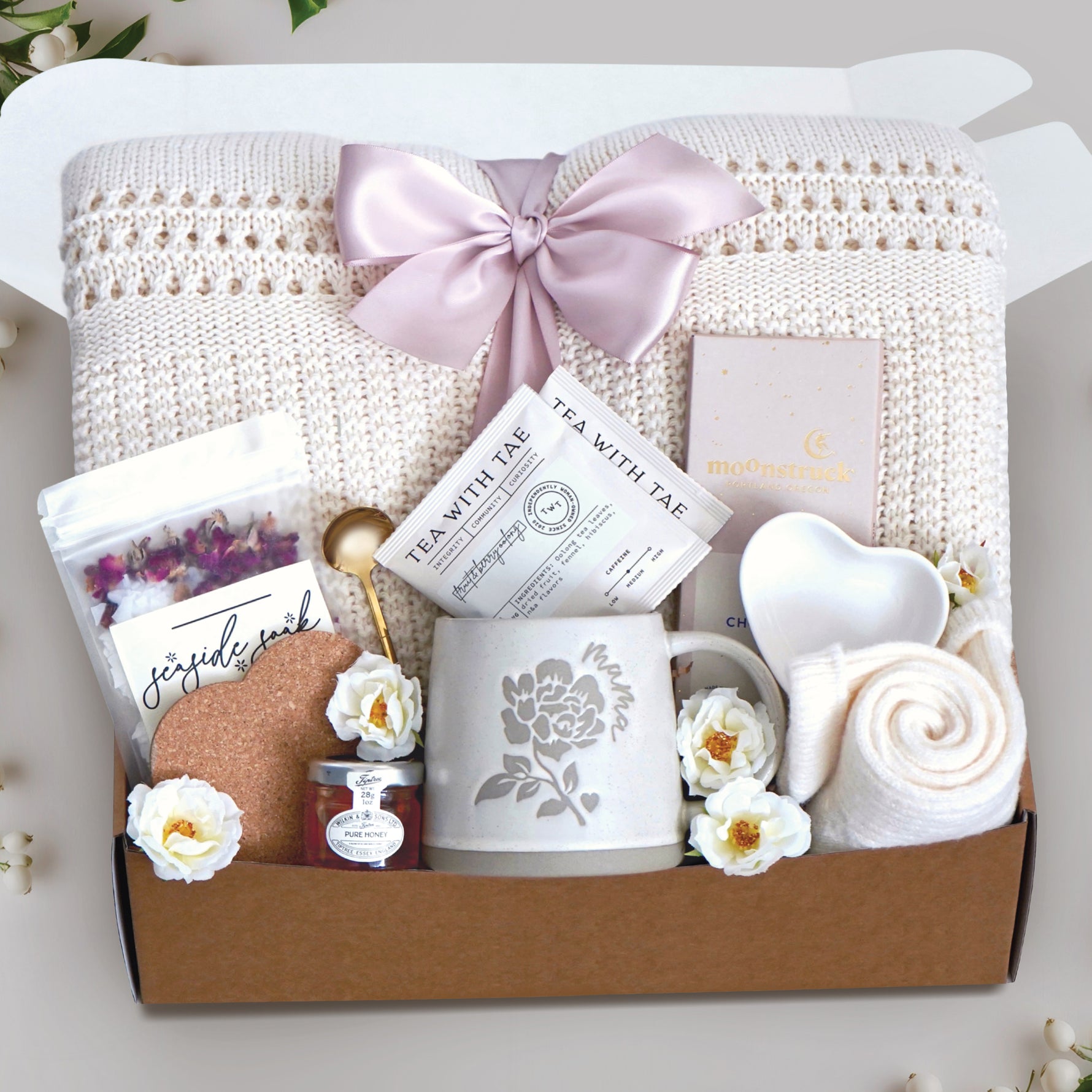 Love you mama blanket box comes with a cream lace knit blanket tied in a blush ribbon, 2 tea with tae tea bags, gold demitasse spoon, rose seaside soak bath salts, heart coaster, mini honey, mama ceramic mug, heart trinket dish, full size moonstruck milk chocolate bar, and cream knit socks