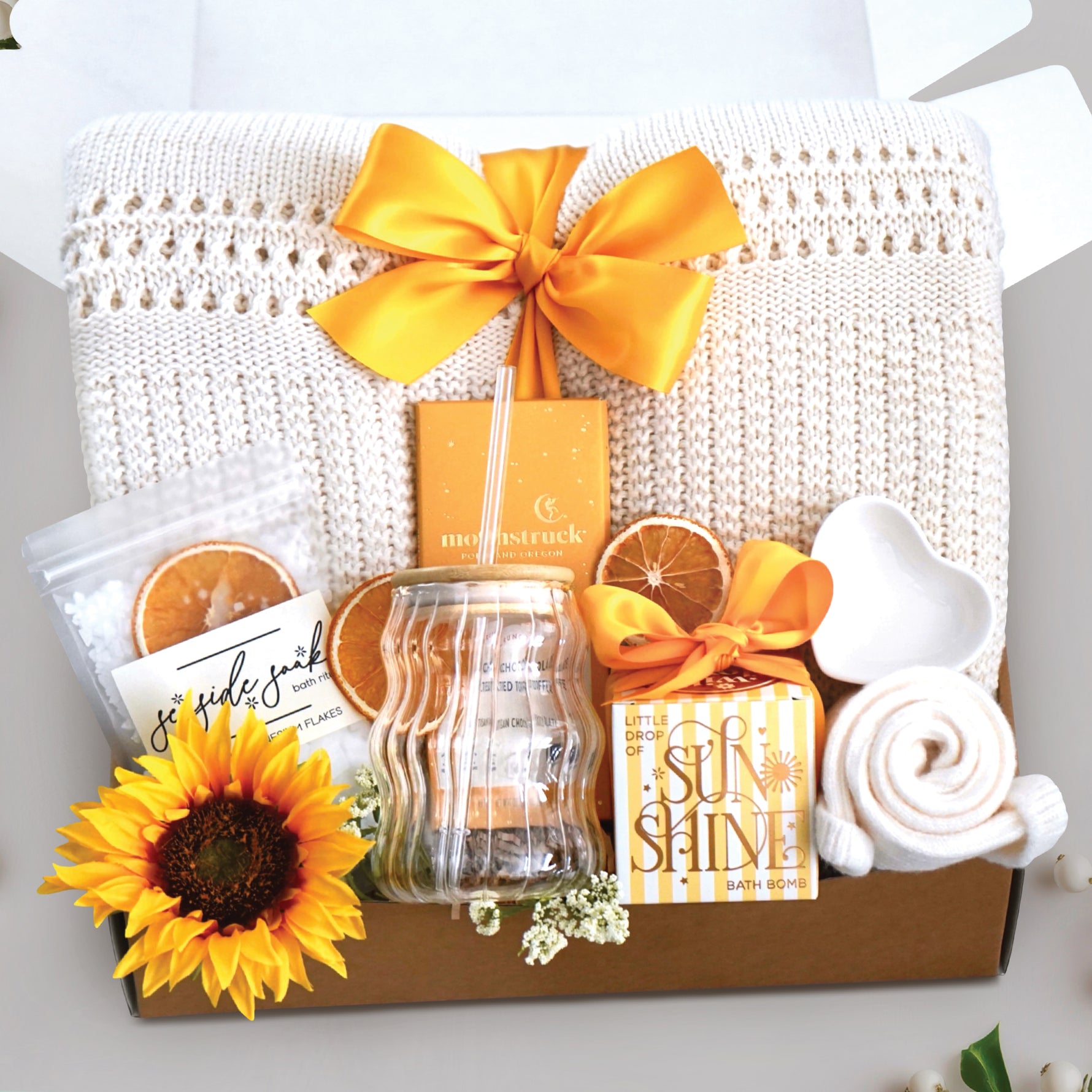 My sunshine blanket box comes with a cream lace knit blanket tied with a yellow ribbon, seaside soak bath salts with orange slices, wavy glass tumbler and straw, full size moonstruck milk chocolate and toffee bar, little drop of sunshine bath bomb, heart trinket tray, and cream knit socks