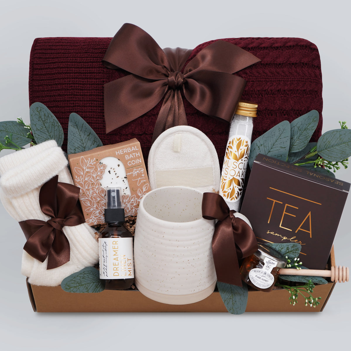 Wildflower Pop Ruby Spa Box with Burgundy blanket, bath salts, bath bomb, linen mist, cream socks, tea sampler, and mini honey and dipper. On light grey background