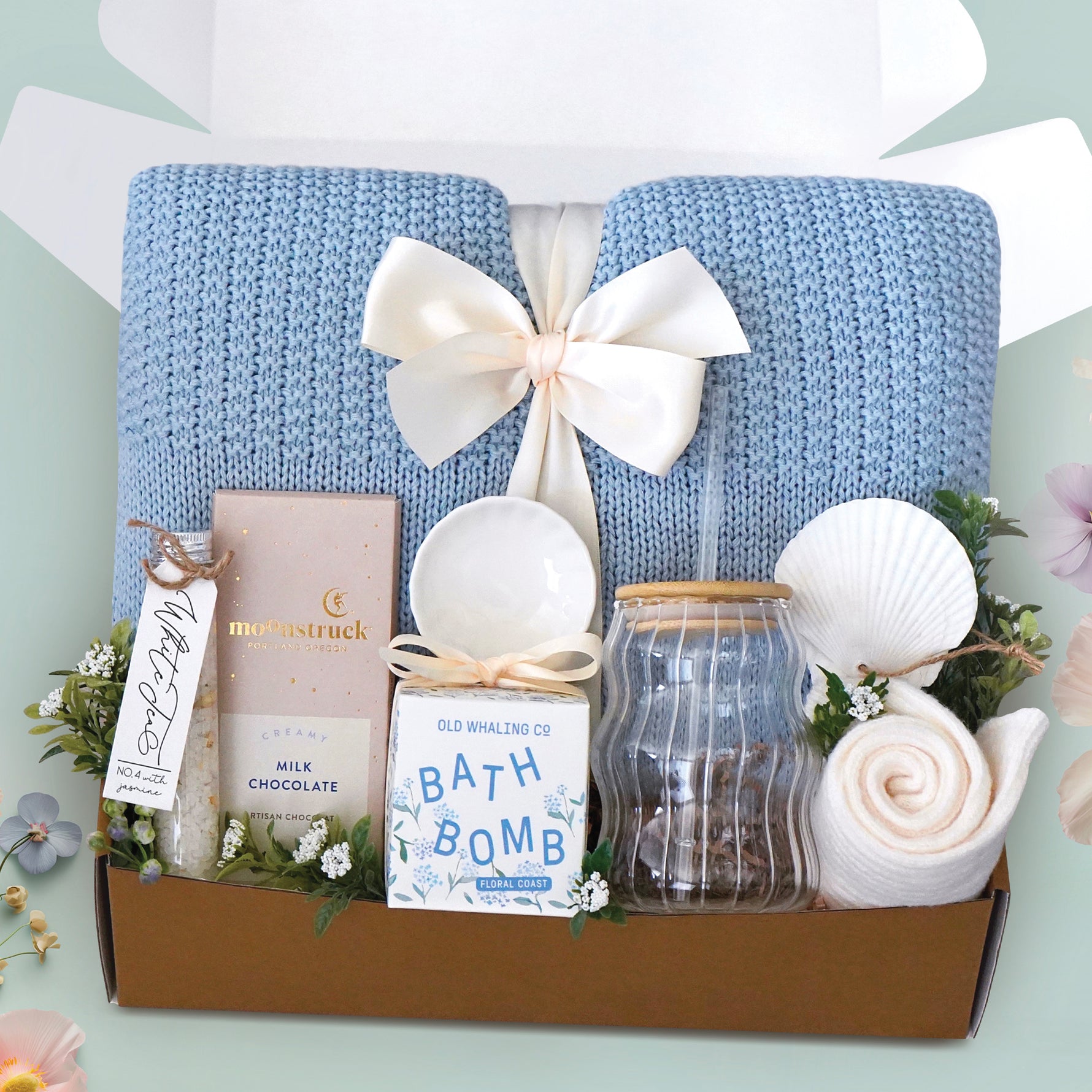 The Sea Glass Blanket Box comes with a blue lace knit blanket tied in a cream ribbon, full size Moonstruck milk chocolate bar, White tea bath salts, Old Whaling co floral coast bath bomb, scallop trinket dish, wave glass tumbler with plastic straw, sea shell ornament, and cream knit socks