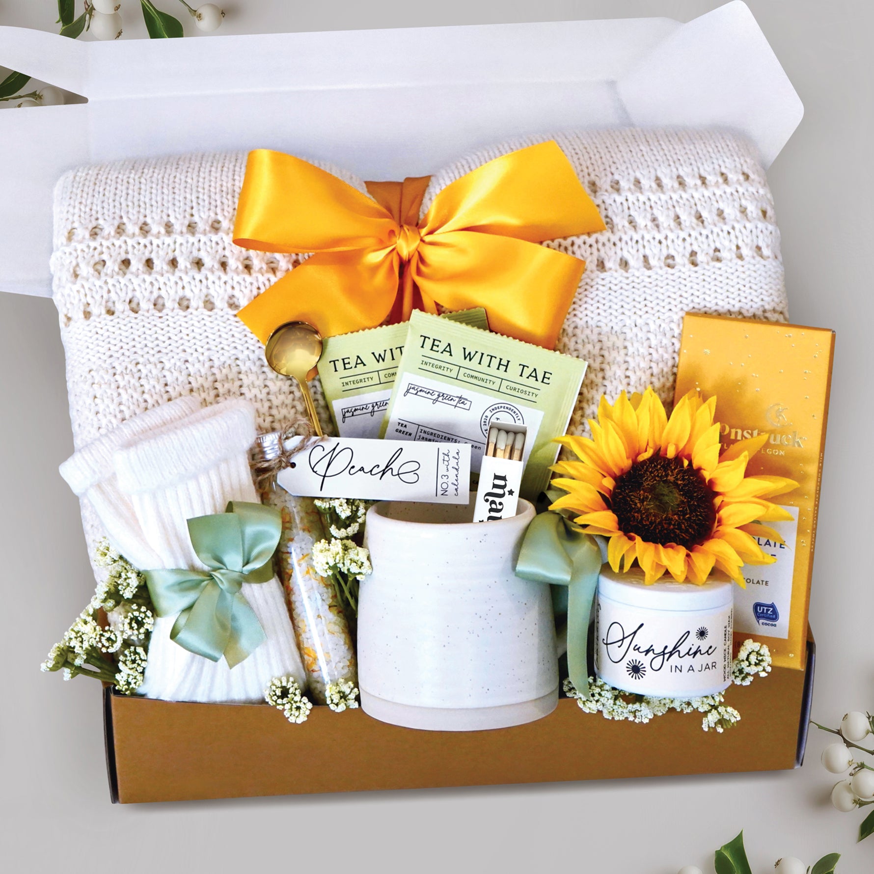 Sending Sunshine Gift Box comes with cream lace knit throw tied with sunshine yellow ribbon, tea sampler, tube of bath salts, trail size soy wax candle and matches, toffee and milk chocolate moonstruck bar, cream knit socks, and faux sunflower head.