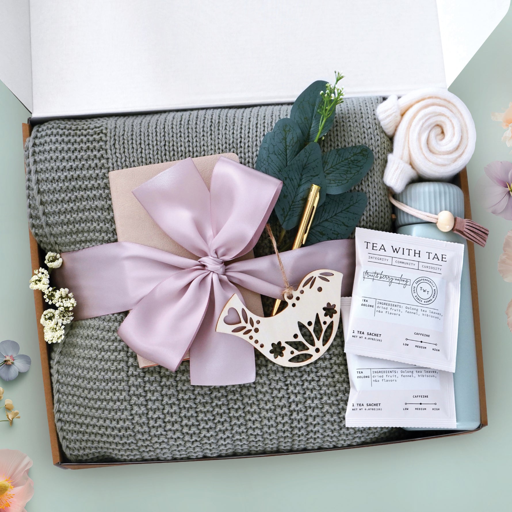 The summer sonnet blanket box comes with a sage green lace knit blanket tied with a blush ribbon, notebook, gold ballpoint pen, bird nordic wood ornament, 2 tea with tae bags, sea foam green insulated tumbler, cream knit socks, and faux eucalyptus sprig