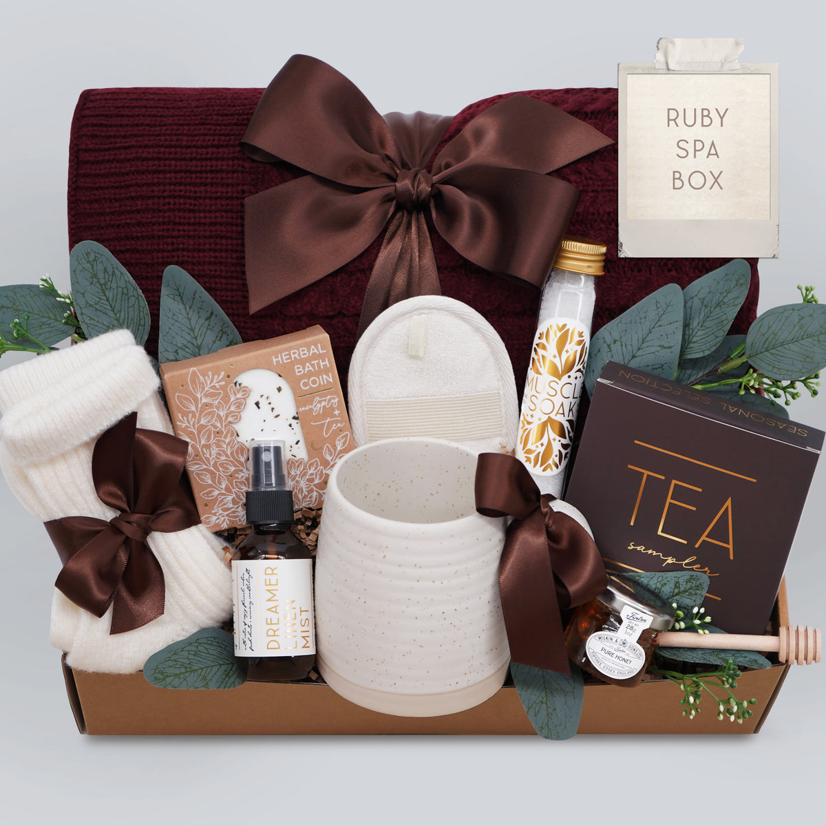 Wildflower Pop Ruby Spa Box with Burgundy blanket, bath salts, bath bomb, linen mist, cream socks, tea sampler, and mini honey and dipper. On light grey background
