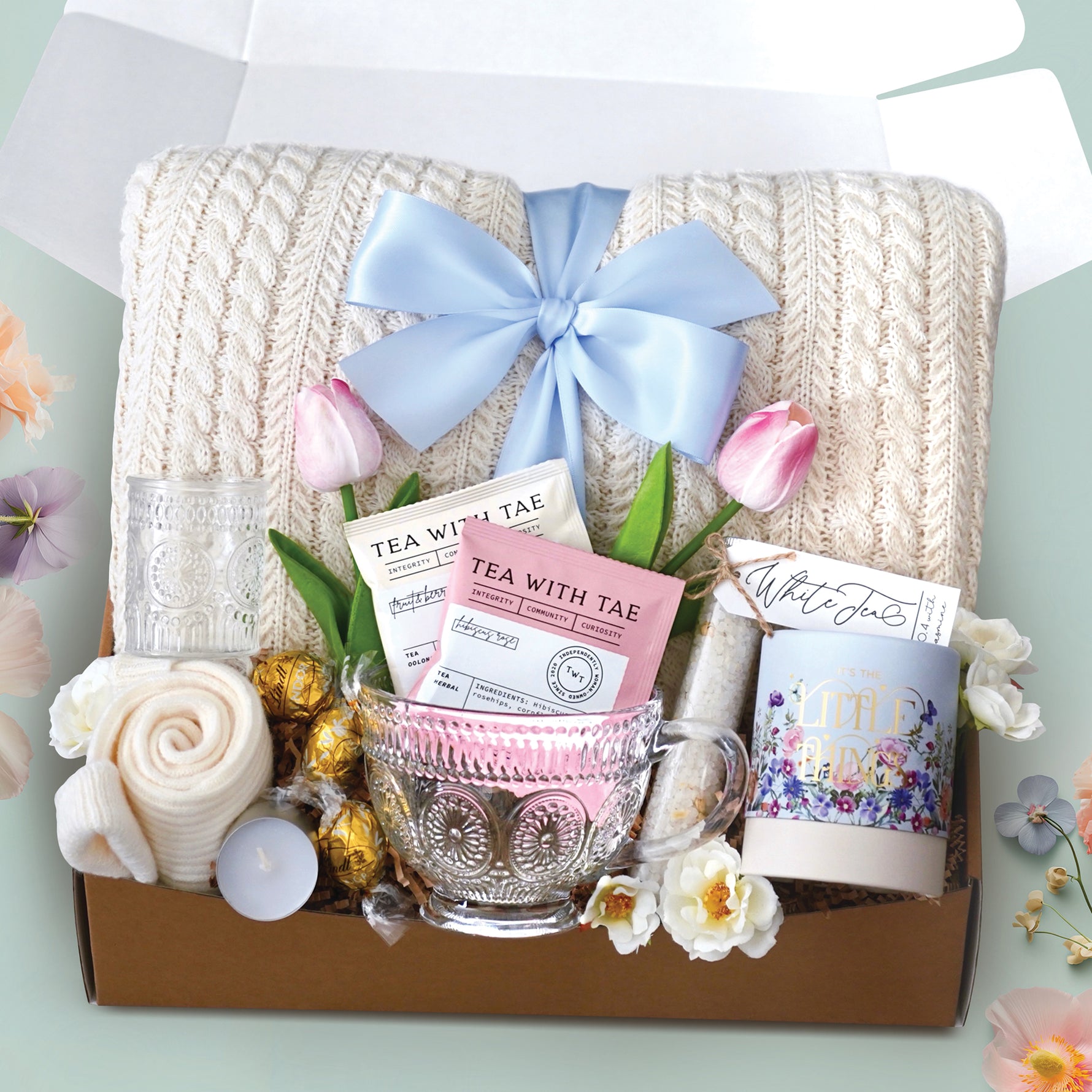 The Tulip time blanket box comes with a cream cable knit blanket tied with a baby blue ribbon, it's the little things truffle tube, bath salt tube, 2 tea bags, glass mug, glass votive and tea light, cream knit socks, and a faux tulip stem