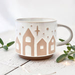 White ceramic mug with starry village design on a light wooden surface with greenery