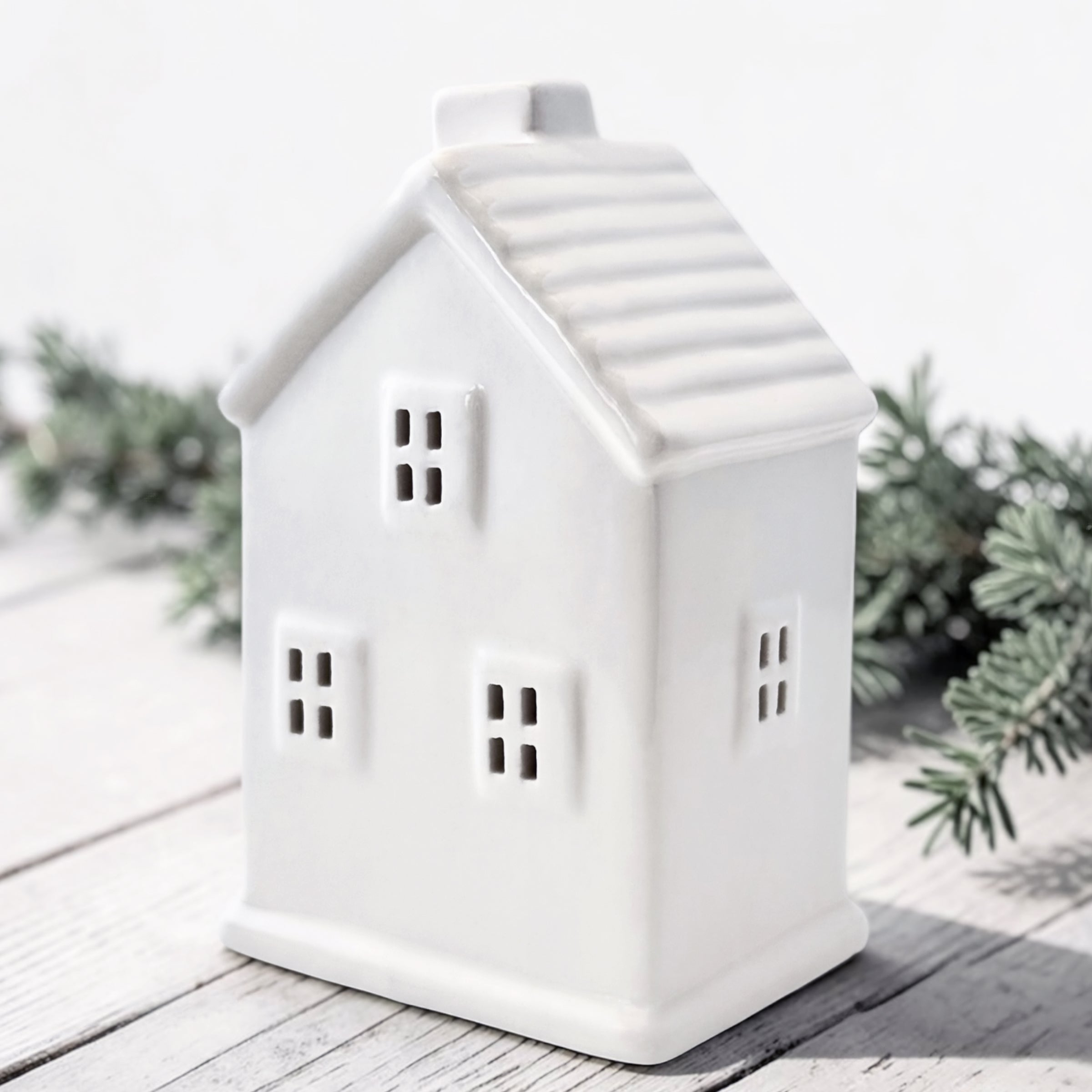 A white ceramic house with window cutouts, photographed on a light wood surface with soft greenery in the background.