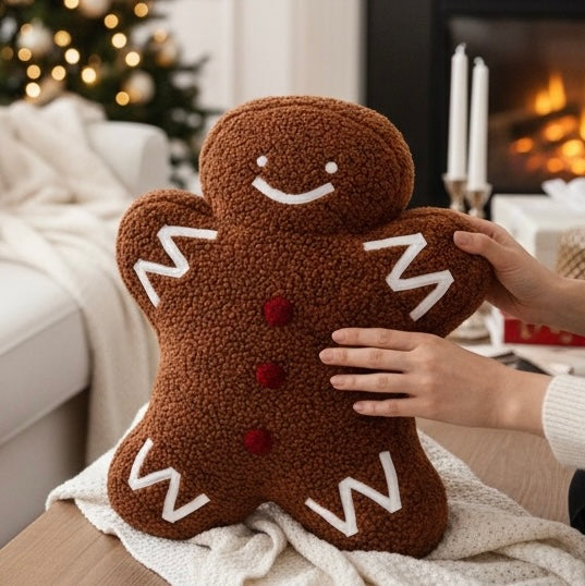 Gingerbread man pillow with white icing details and red buttons, being held by hands in a cozy living room setting with a fireplace and Christmas tree in the background.