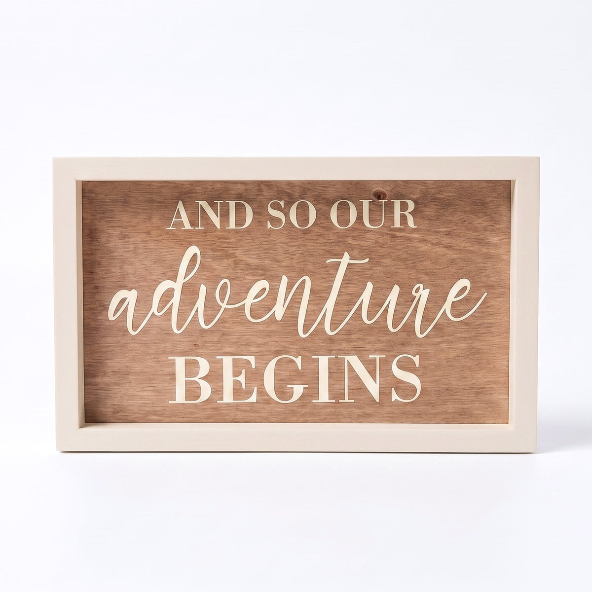 A rectangular wooden sign with a light painted frame and a medium brown wood-grain center panel. The text reads 'AND SO OUR ADVENTURE BEGINS' in cream-colored lettering, with 'adventure' in a cursive script font.