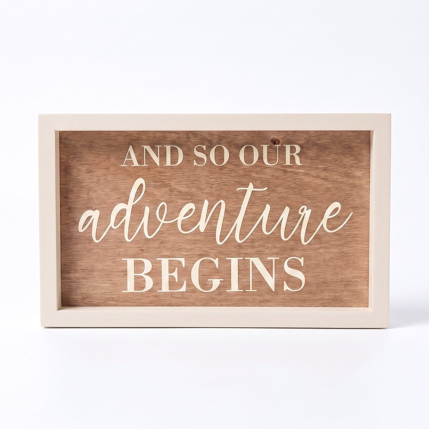 A rectangular wooden sign with a light painted frame and a medium brown wood-grain center panel. The text reads 'AND SO OUR ADVENTURE BEGINS' in cream-colored lettering, with 'adventure' in a cursive script font.