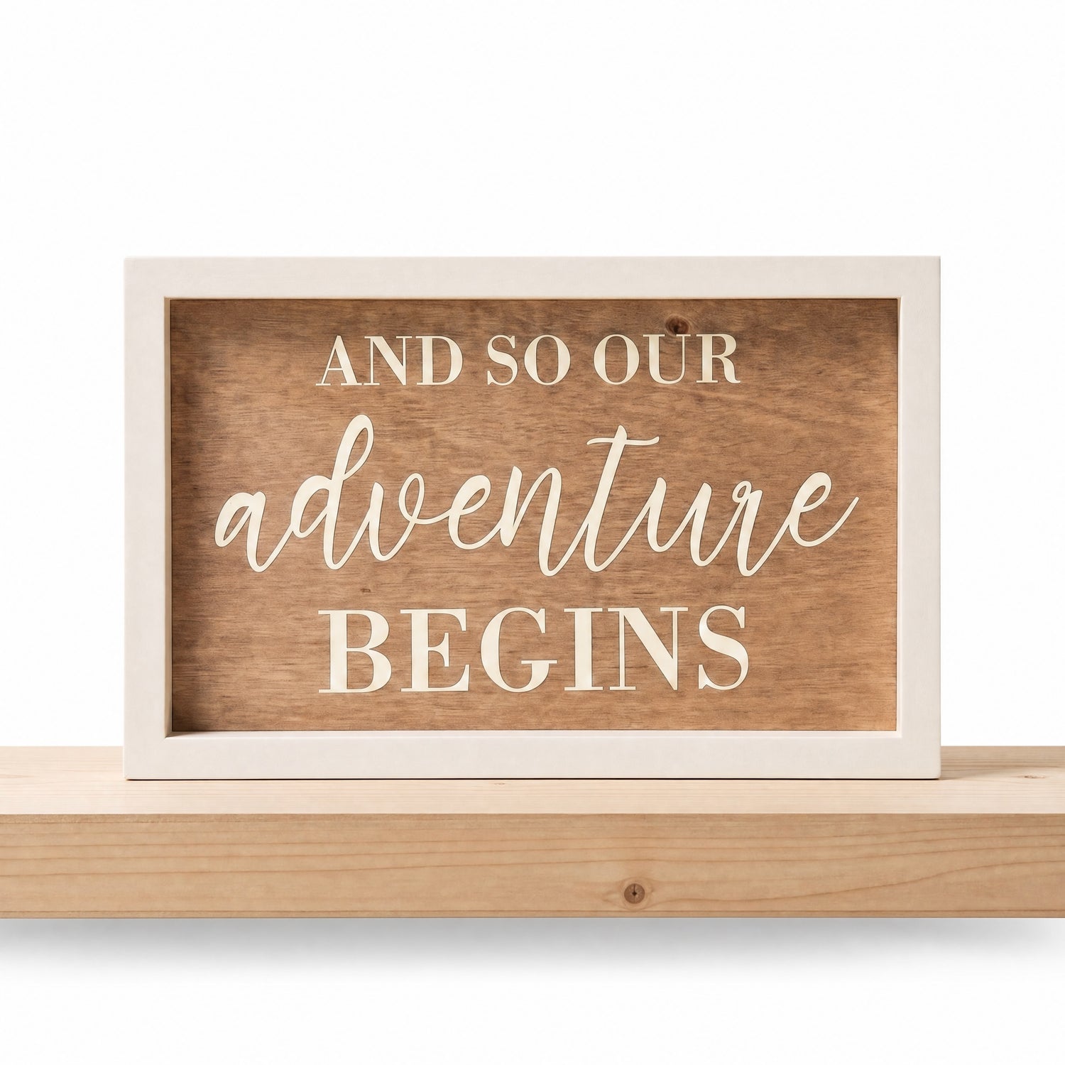 A rectangular wooden sign with a light painted frame and a medium brown wood-grain center panel. The text reads 'AND SO OUR ADVENTURE BEGINS' in cream-colored lettering, with 'adventure' in a cursive script font. Sign is sitting on a neutral wood shelf, not included.