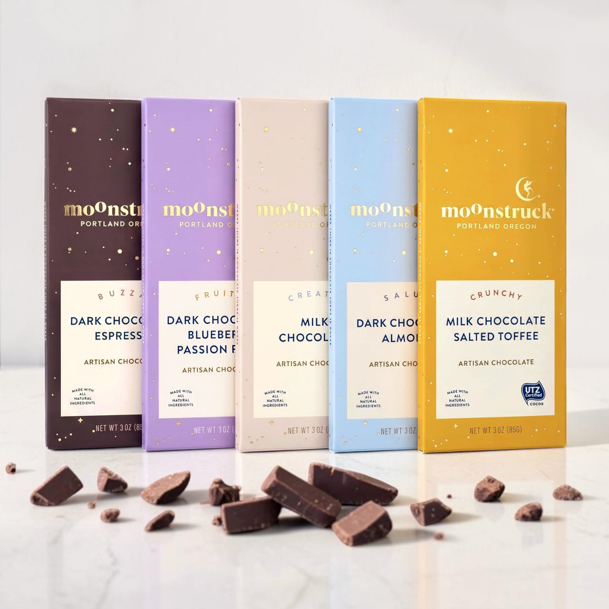 Moonstruck artisan chocolate bars in different flavors lined up on a white surface with scattered chocolate pieces in front.

Flavors from left to right, dark chocolate espresso, dark chocolate blueberry passion fruit, milk chocolate, dark chocolate almond, milk chocolate salted toffee. 