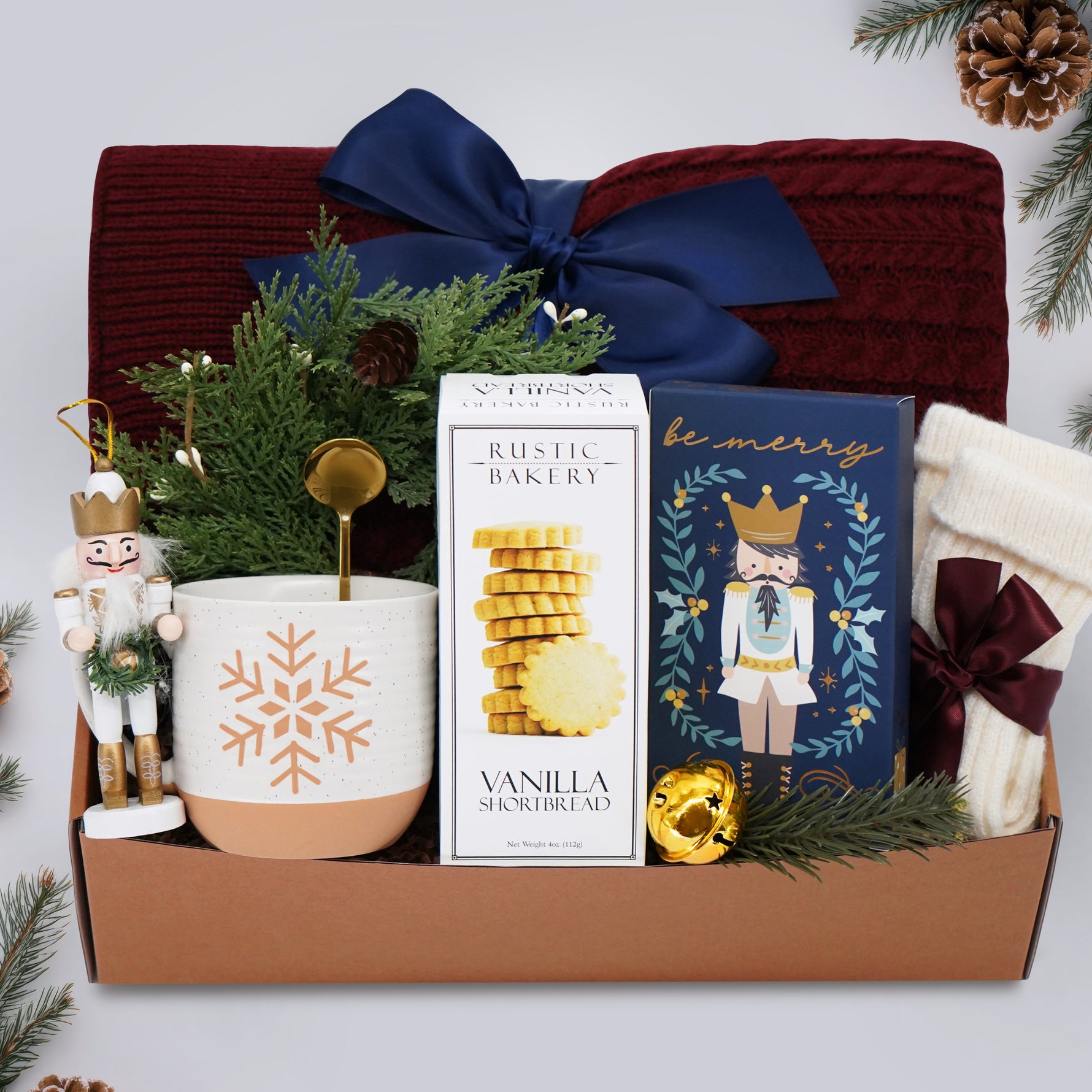 Wildflower Pop Be Merry Gift box with Burgundy cable knit blanket, shortbread cookies, snowflake mug, nutcracker ornament, be merry drink sampler, mini wreath, cream knit socks, gold demitasse spoon, and gold jingle bell on a grey background