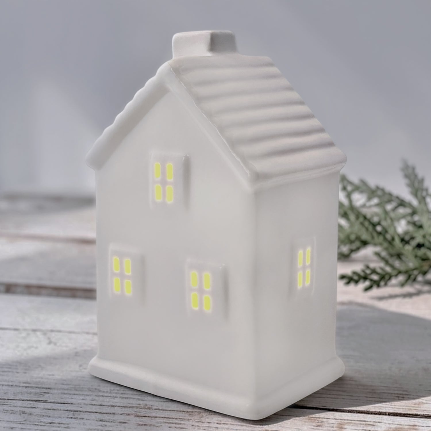 A white ceramic house with window cutouts, led light inside, photographed on a light wood surface with soft greenery in the background.