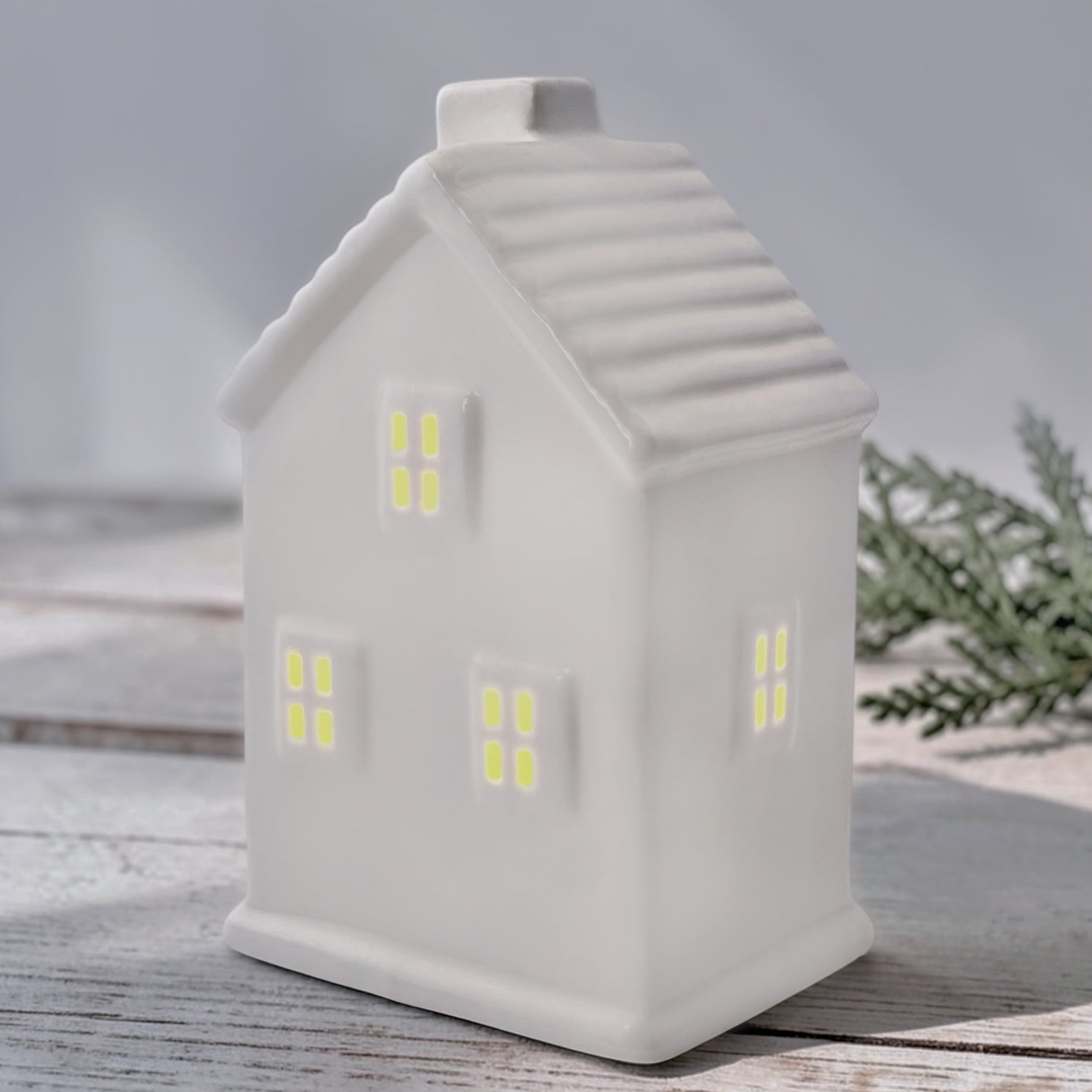 A white ceramic house with window cutouts, led light inside, photographed on a light wood surface with soft greenery in the background.
