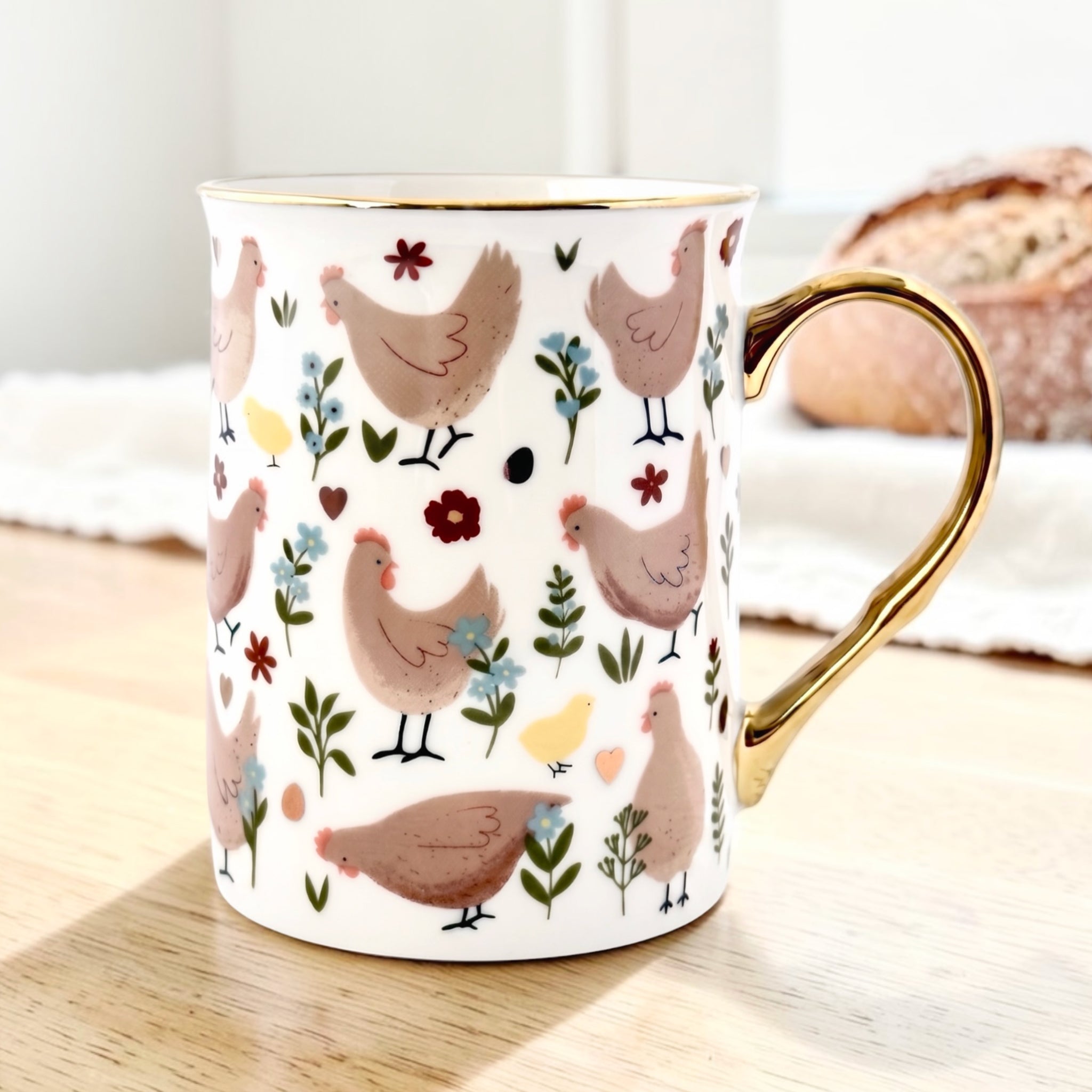 A white ceramic mug with a gold handle and rim, decorated with a pattern of brown chickens and small colorful flowers, sitting on a wooden table.