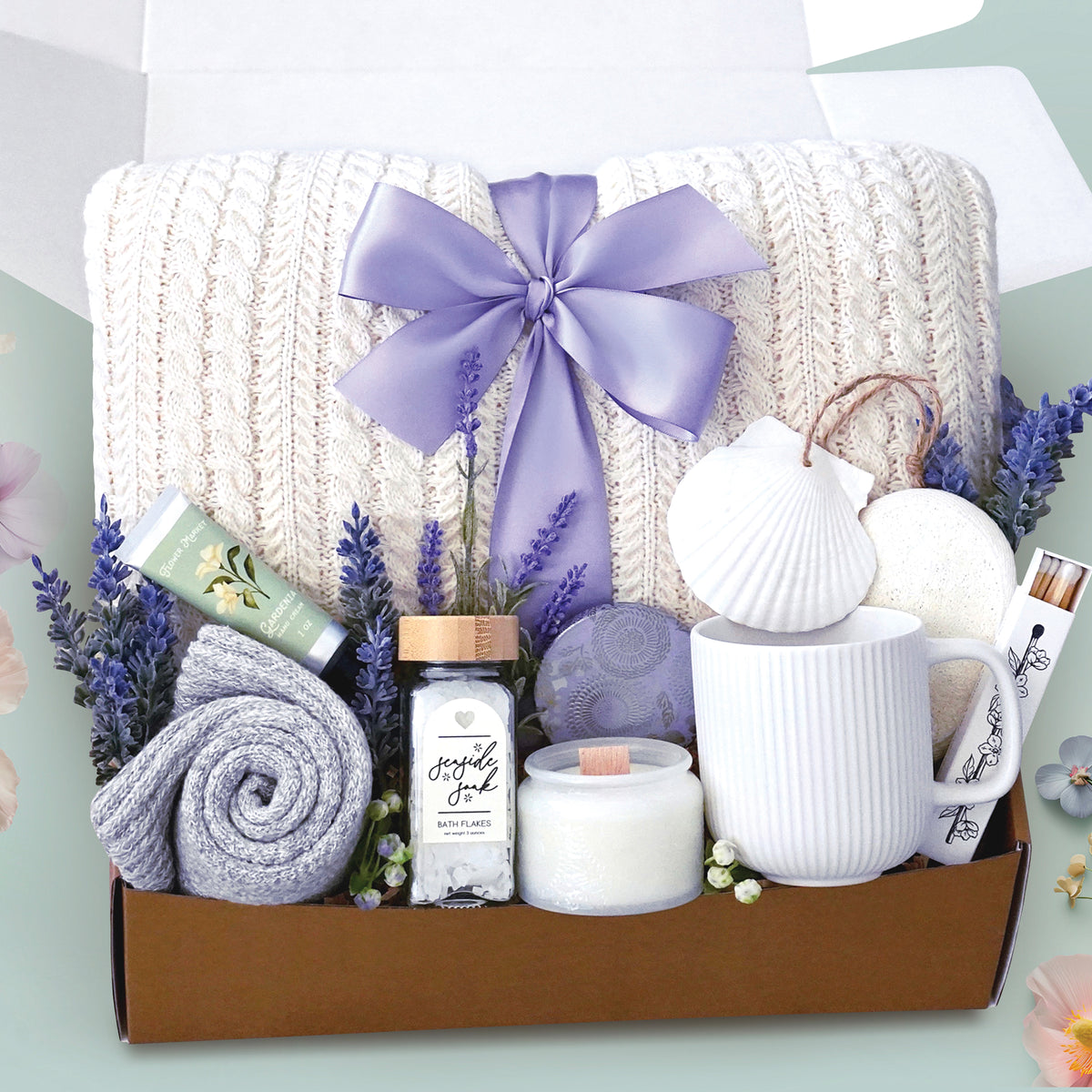 The Costal Cottage comes with a cream cable knit blanket tied with a lavender bow, a white ceramic mug, cozy boot socks, seaside bath soak, Gardenia hand cream, a soy wax candle, box of matches, and exfoliating sponge, and a seashell ornament. Package with care in a branded wildflower pop box and sent to your loved one.  