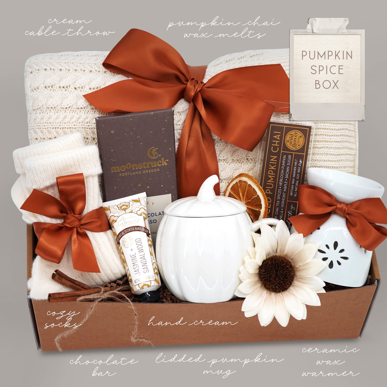 Wildflower Pop Pumpkin Spice Blanket Gift Box, cream cable knit blanket with pumpkin orange ribbon, chocolate, cream socks, cream pumpkin mug with lid, hand lotion, wax warmer and wax melt. On a grey background