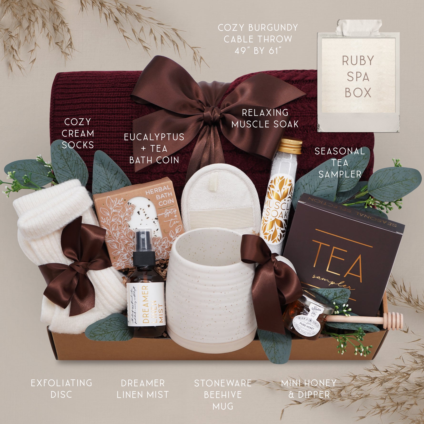 Wildflower Pop Ruby Spa Box with Burgundy blanket, bath salts, bath bomb, linen mist, cream socks, tea sampler, and mini honey and dipper. On tan grey background 