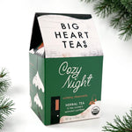 A box of Big Heart Teas Cozy Night herbal tea sachets, styled with pine branches on a white background.