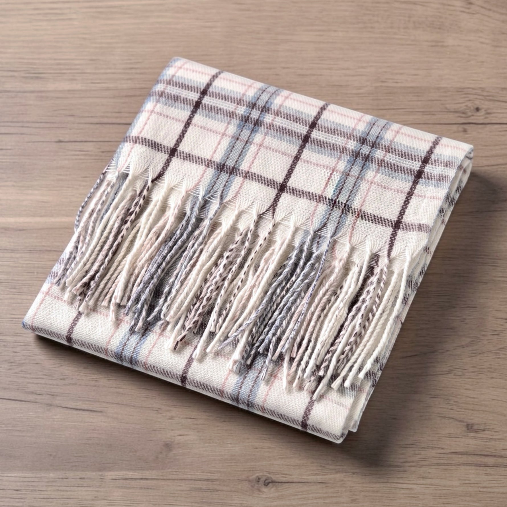 A folded cream plaid scarf with fringe, displayed on a light brown wooden surface. The scarf features a cream base with a classic plaid pattern in coordinating shades of dark brown, dusty blue, and rust.