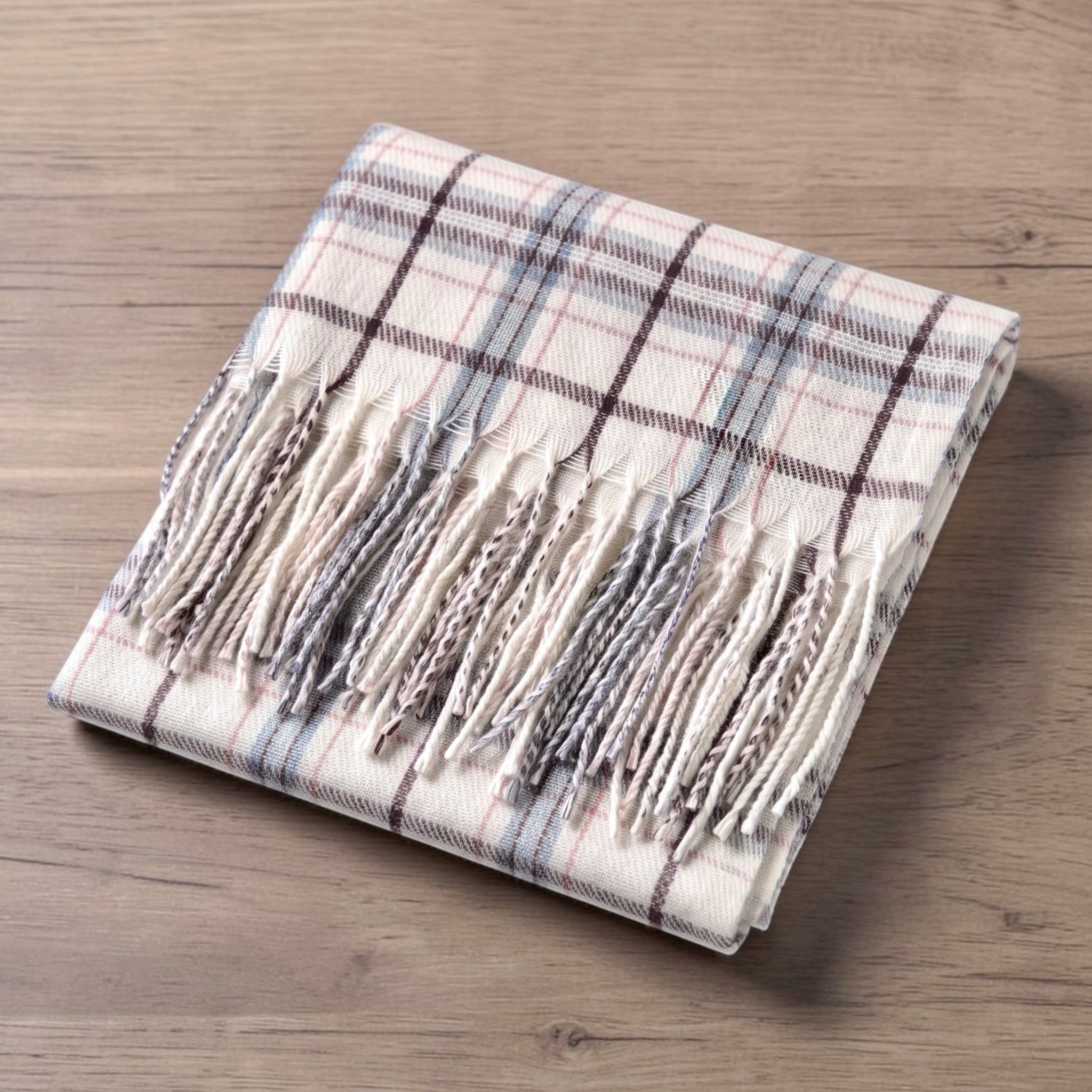 A folded cream plaid scarf with fringe, displayed on a light brown wooden surface. The scarf features a cream base with a classic plaid pattern in coordinating shades of dark brown, dusty blue, and rust.