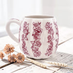 A white ceramic pumpkin mug with a red floral pattern, styled on a rustic wooden surface with dried flowers and wheat stalks.