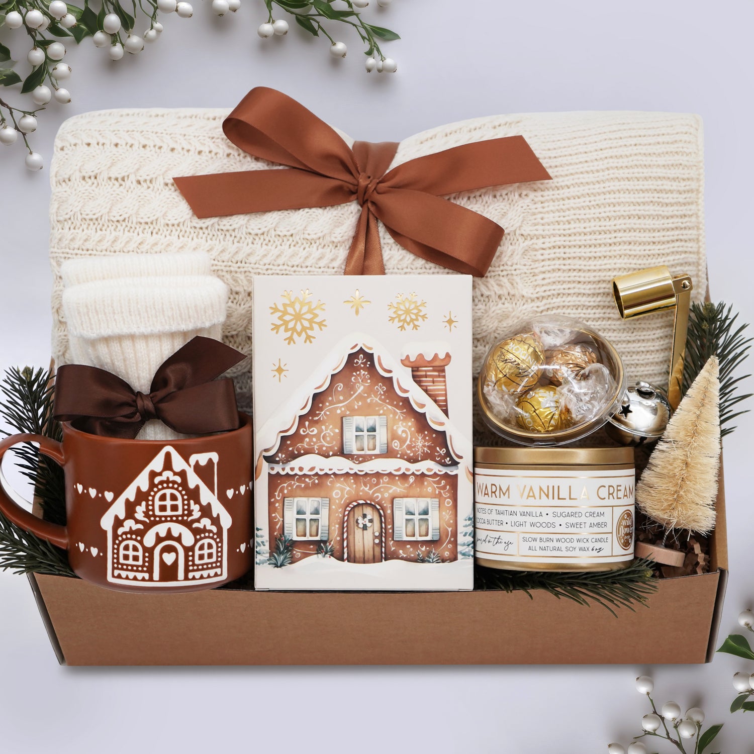 Wildflower Pop Gingerbread Cottage Blanket gift box includes a cream cable knit blanket tied with a ginger ribbon, Wildflower Pop exclusive gingerbread Mug, cream knit socks, truffle dome sampler, 8 oz poured in the USA soy wax candle in vanilla scent, gold candle snuffer, cream mini tree, and silver jingle bell