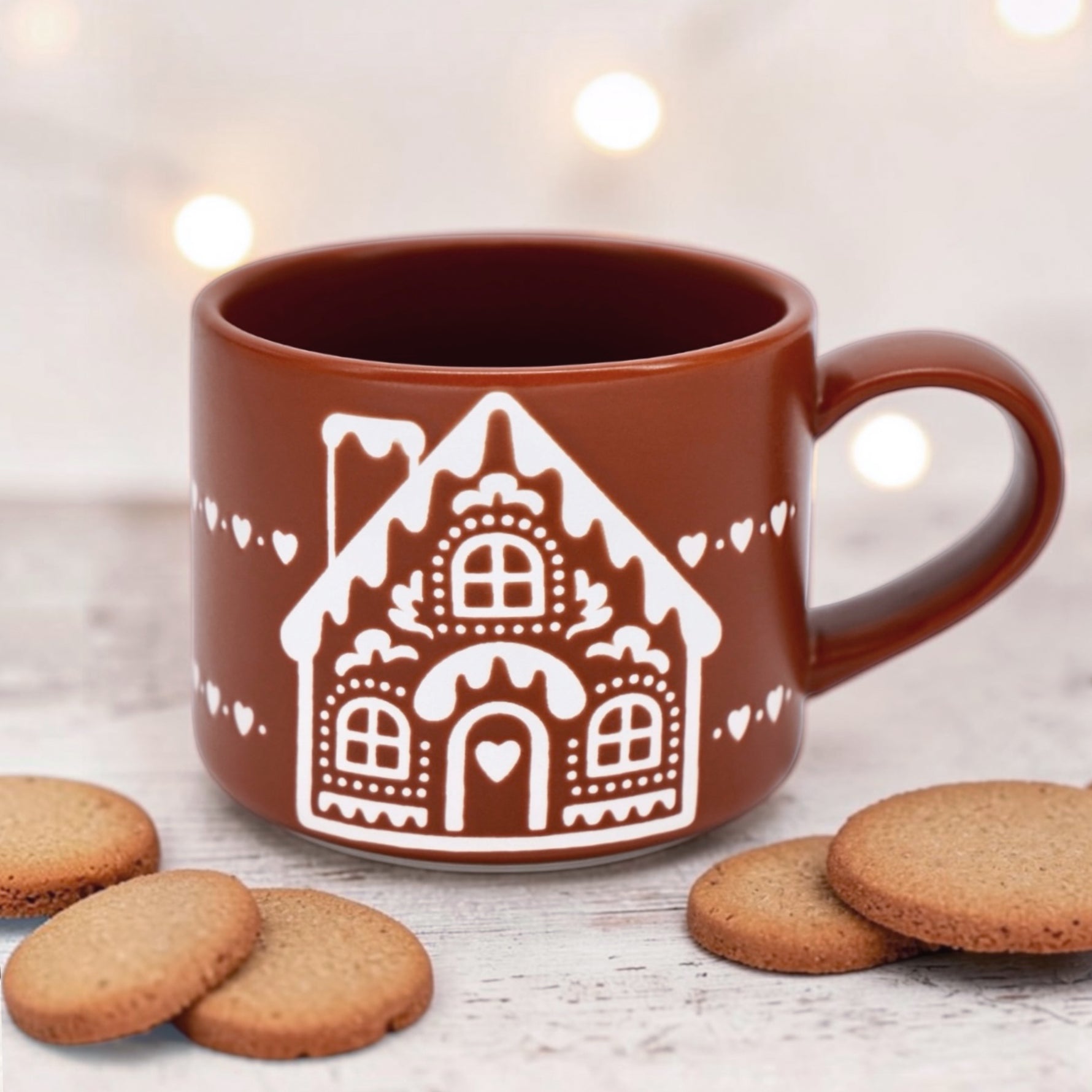 A gingerbread colored ceramic mug with a white gingerbread house design, surrounded by cookies on a light wooden surface with warm bokeh lights in the background