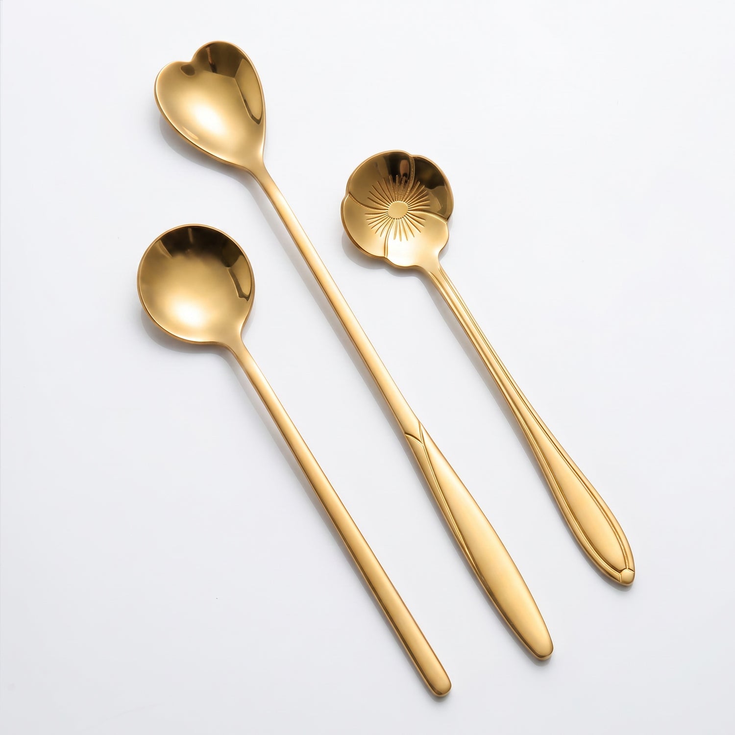 Three gold-colored metal spoons arranged diagonally on a clean white background. One spoon is heart-shaped, one is flower shaped, and one is a standard round spoon.