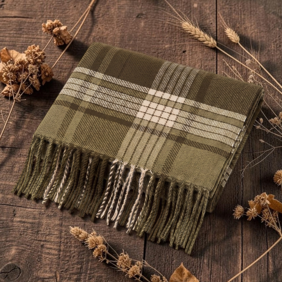 An olive green plaid blanket scarf with cream tones and fringed edges, styled on a rustic dark wood surface with dried flowers.