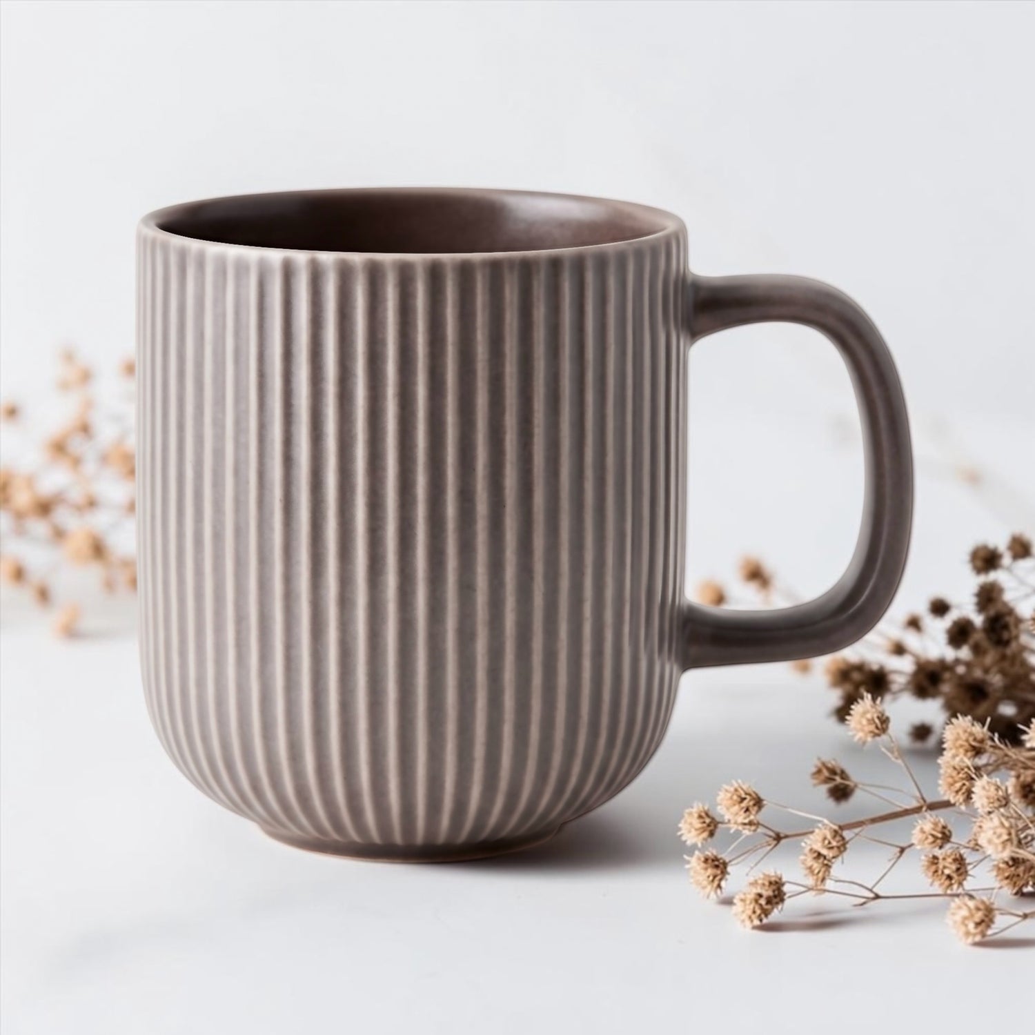 A ribbed ceramic mug in a warm taupe-brown color, styled with dried flowers on a clean white surface.
