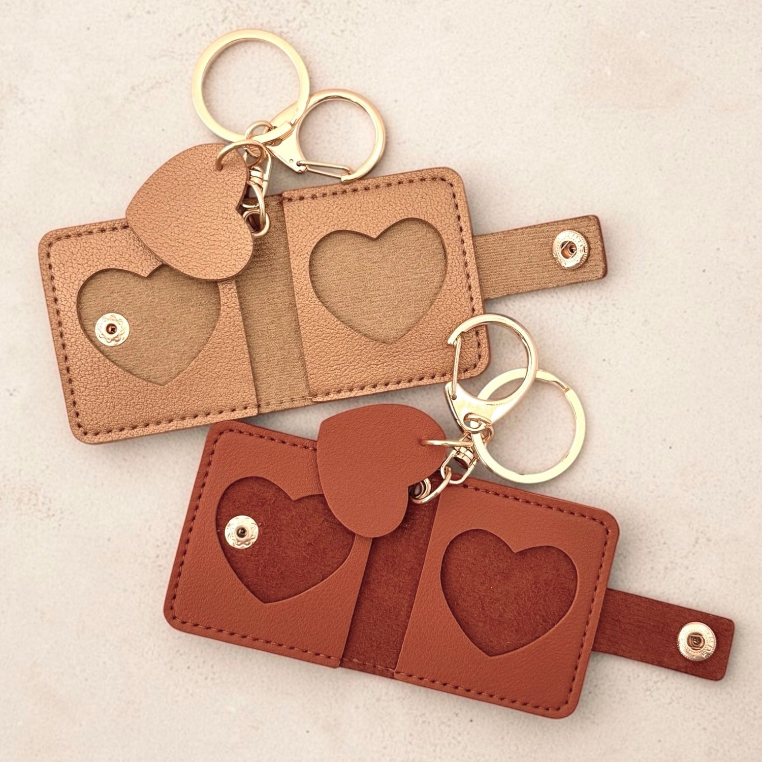 Small leather keychains with heart-shaped cutout picture frames and gold-tone key rings, shown on a light neutral background. Top is gold, bottom is reddish brown.