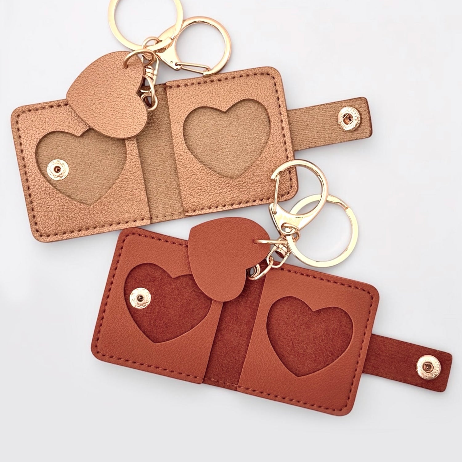 Small leather keychains with heart-shaped cutout picture frames and gold-tone key rings, shown on a light neutral background. Top is gold, bottom is reddish brown.