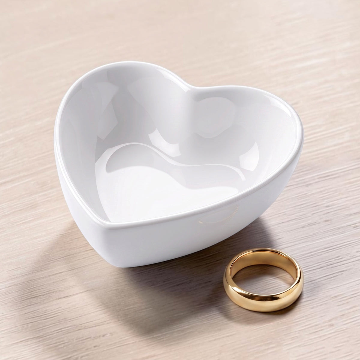 A white heart-shaped ceramic trinket dish placed on a light wood surface, with a gold ring beside it, ring not included.