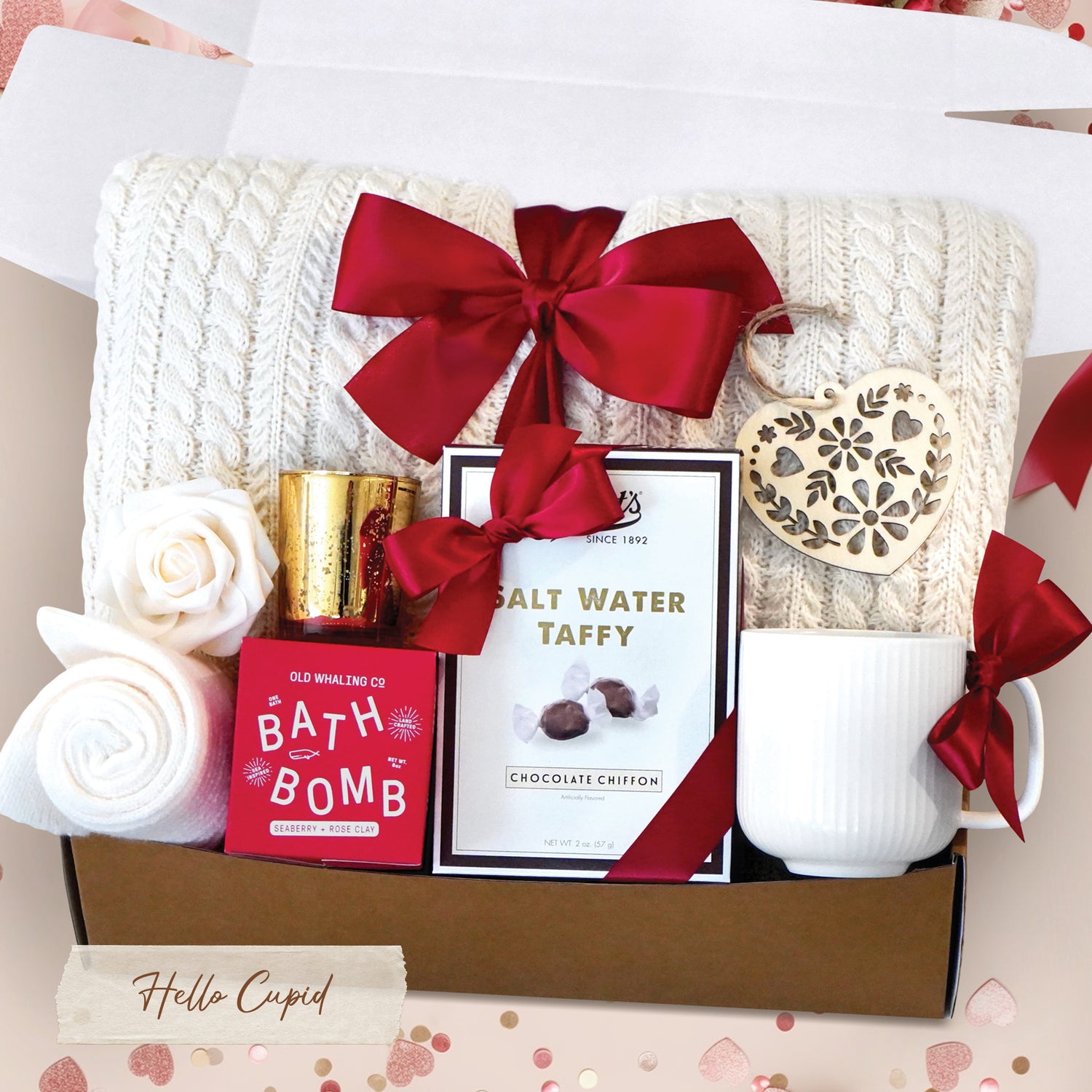Hello Cupid Gift Box comes with a cream cable knit blanket tied with a red ribbon, cream knit socks, gold votive and tea light, Seaberry and Rose bath bomb, Chocolate Chiffon salt water taffy, nordic heart wood ornament, faux rose head, and cream ribbed mug