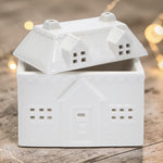A white ceramic house-shaped wax warmer, photographed on a rustic wooden surface with warm blurred fairy lights in the background.