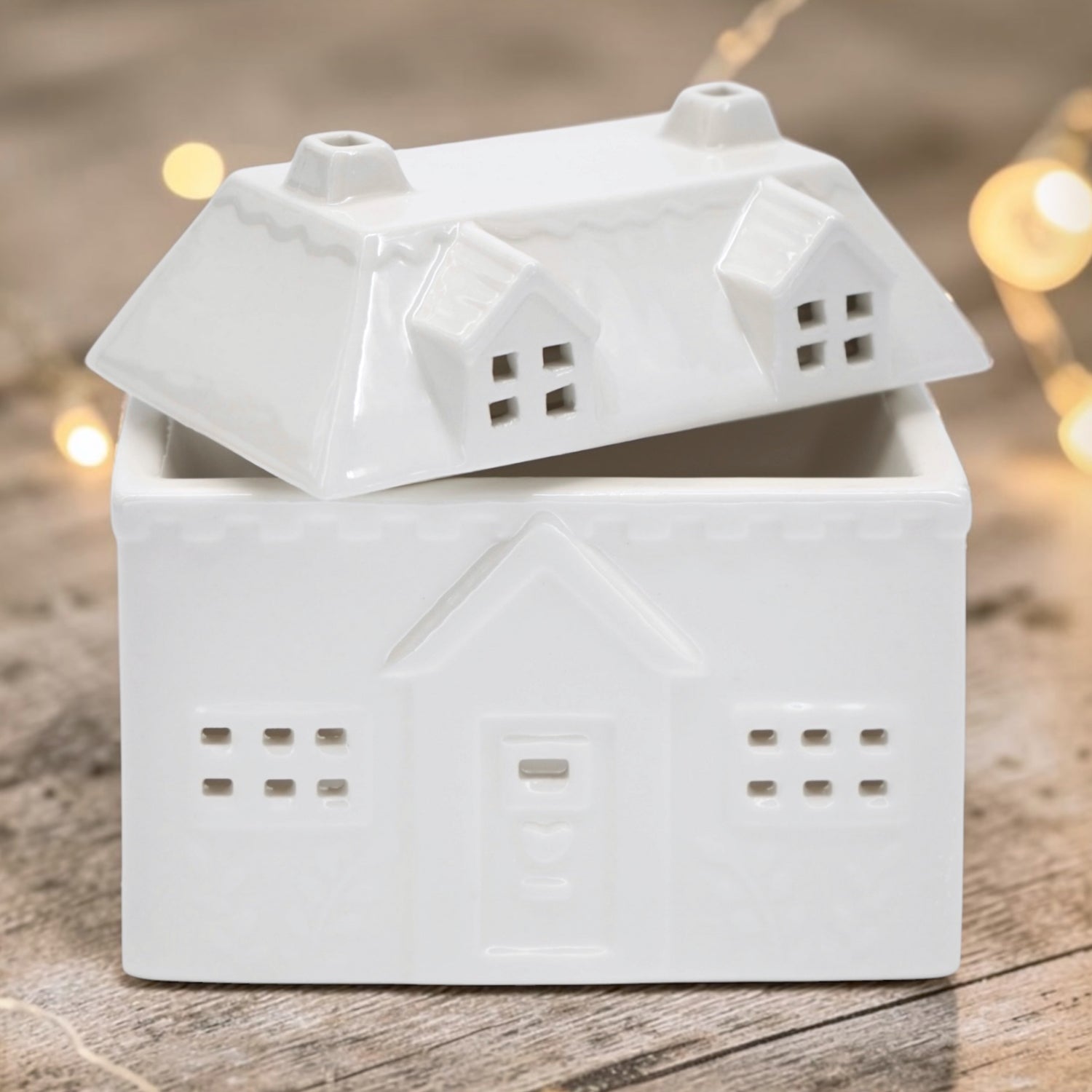 A white ceramic house-shaped wax warmer, photographed on a rustic wooden surface with warm blurred fairy lights in the background.