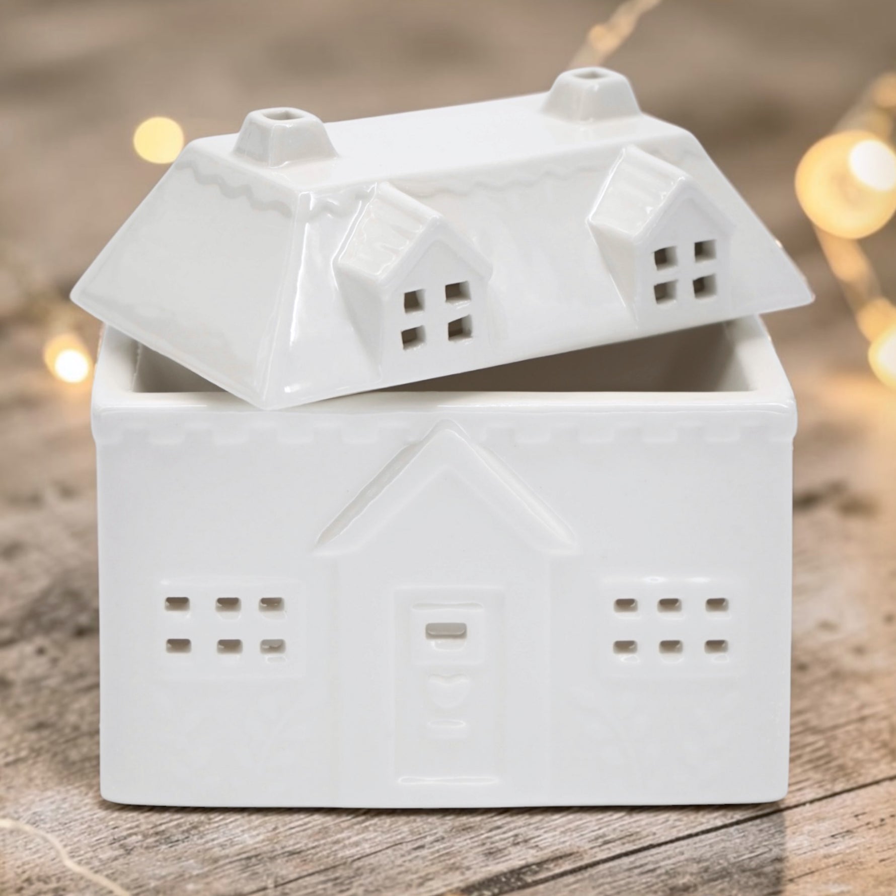 A white ceramic house-shaped wax warmer, photographed on a rustic wooden surface with warm blurred fairy lights in the background.