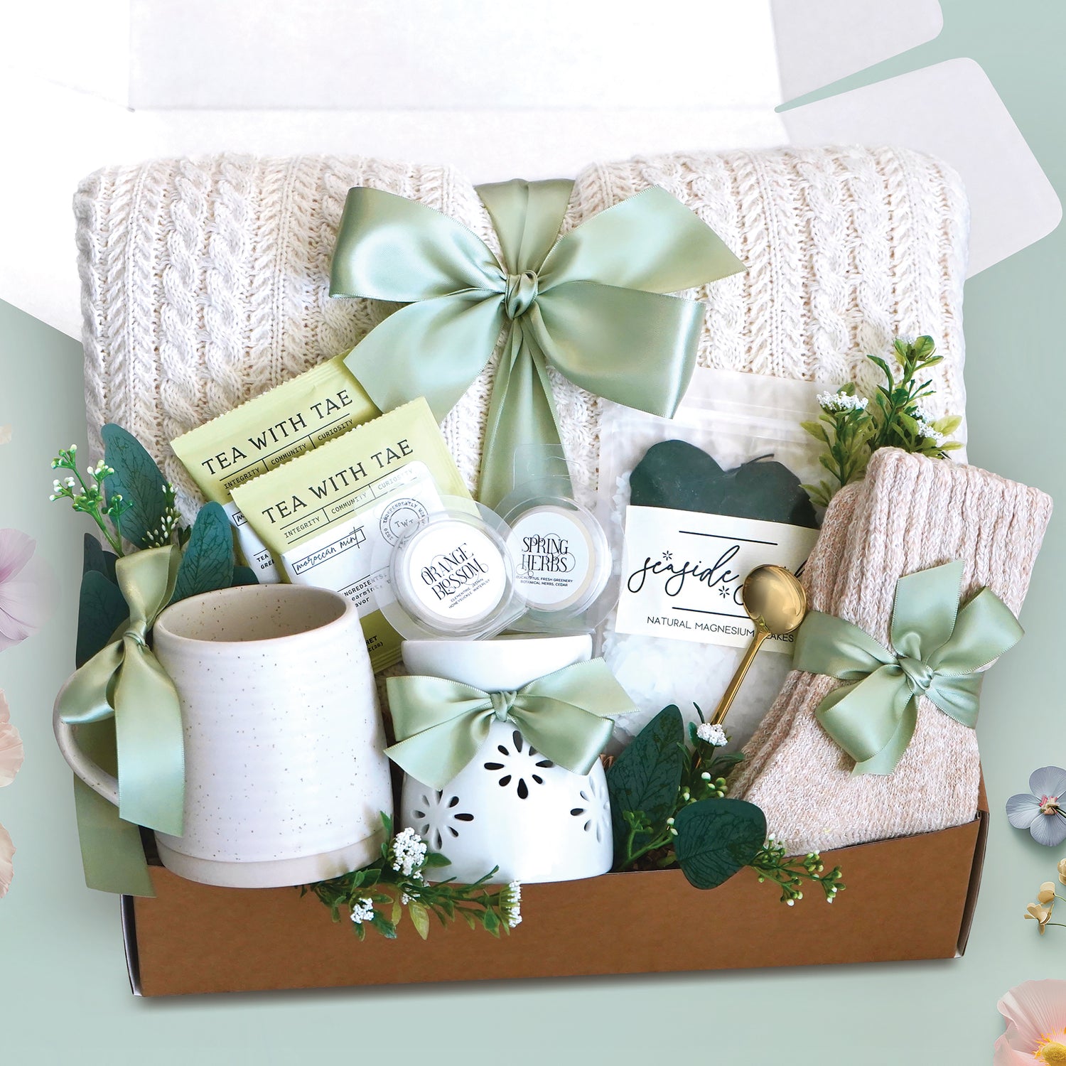The Keep on Growing Gift Box xome with a cream cable knit blanket tied with a sage ribbon, Wildflower Pop Beehive Mug, Starburst wax warmer and 2 wax melts, 2 tea bags, Seaside Soak bath salts, cozy boot socks, and gold demitasse spoon