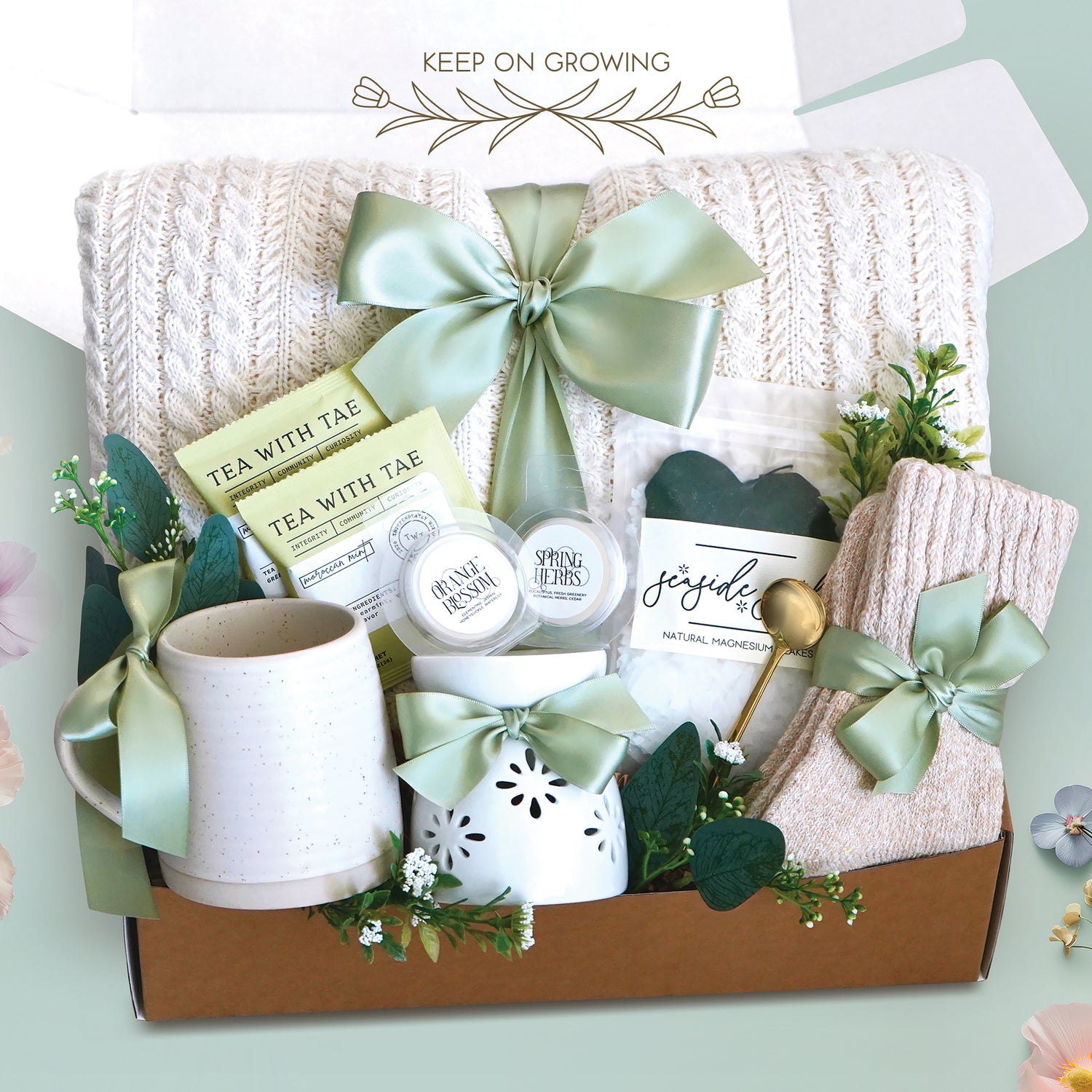 The Keep on Growing Gift Box xome with a cream cable knit blanket tied with a sage ribbon, Wildflower Pop Beehive Mug, Starburst wax warmer and 2 wax melts, 2 tea bags, Seaside Soak bath salts, cozy boot socks, and gold demitasse spoon